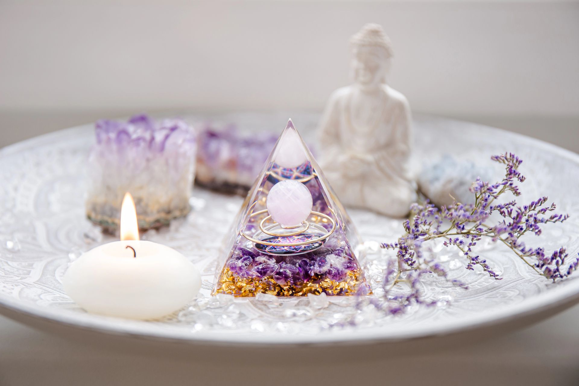 Su un piatto bianco sono appoggiate una piramide di orgone, una statuetta di Buddha, un gruppo di ametiste, una candela bianca e fiori secchi viola.