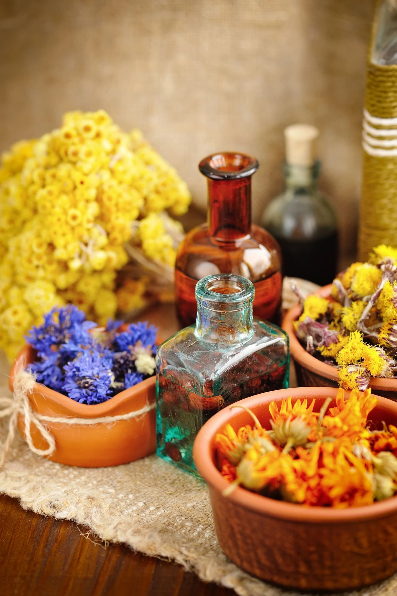 Bottiglie di vetro contenenti oli essenziali, disposte tra ciotole di fiori secchi gialli, arancioni e blu su un panno dalla trama particolare.