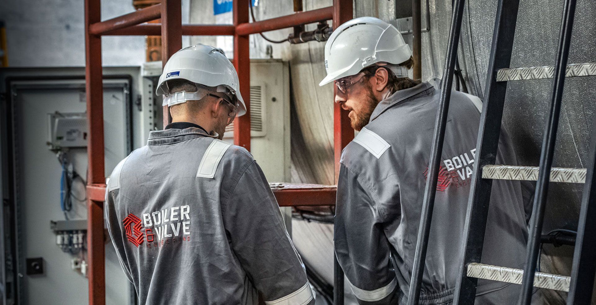 Two workers in gray uniforms and hard hats inspecting equipment at a work site