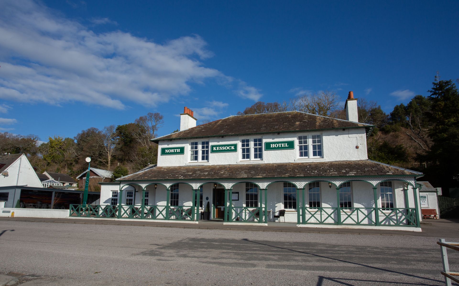 White two-story inn with green trim and porch under a blue sky, seen from across a gravel lot