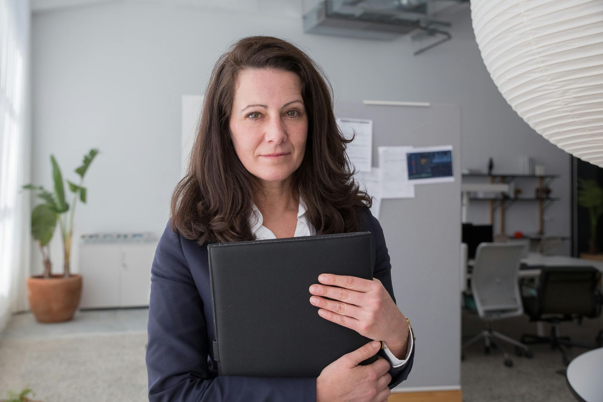 Woman holding a black folder in a modern office, standing beside a desk with a neutral expression.