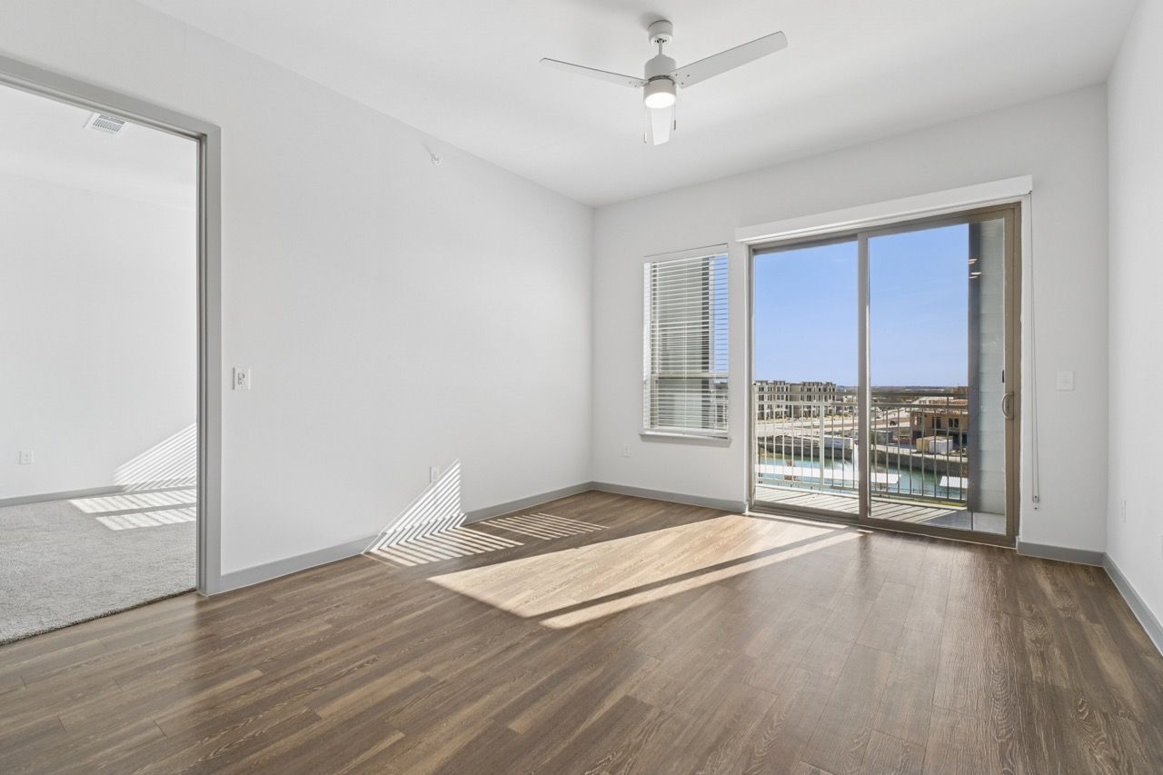 Empty living room with wood-look flooring, white walls, a ceiling fan, and a sliding door to the balcony  at Ennova Fulshear in Fulshear, TX.