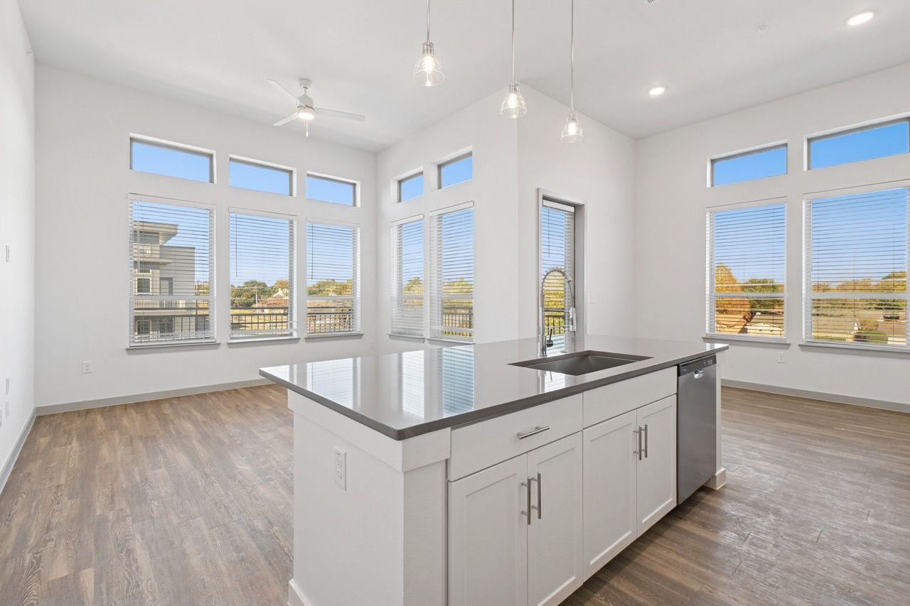 Bright open kitchen with white cabinets, a large central island, and a stainless steel sink beneath expansive windows  at Ennova Fulshear in Fulshear, TX.