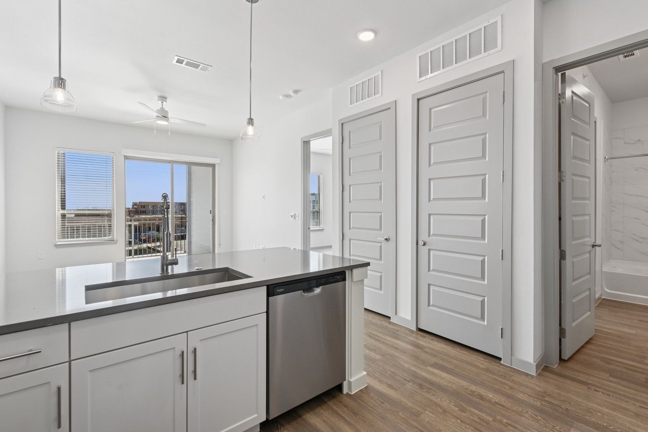Open-concept kitchen in a bright apartment with an island, stainless dishwasher, and balcony door  at Ennova Fulshear in Fulshear, TX.