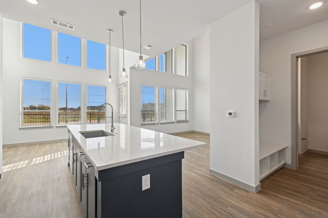Open-concept kitchen with a large white island, sink, pendant lights, and tall bright windows at Ennova Fulshear in Fulshear, TX.