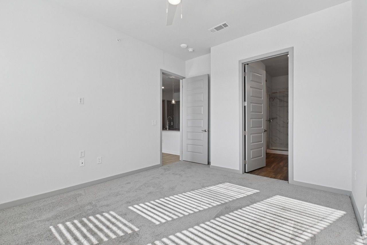 Empty bedroom with white walls, gray carpet, and two doors to a bathroom and hallway  at Ennova Fulshear in Fulshear, TX.