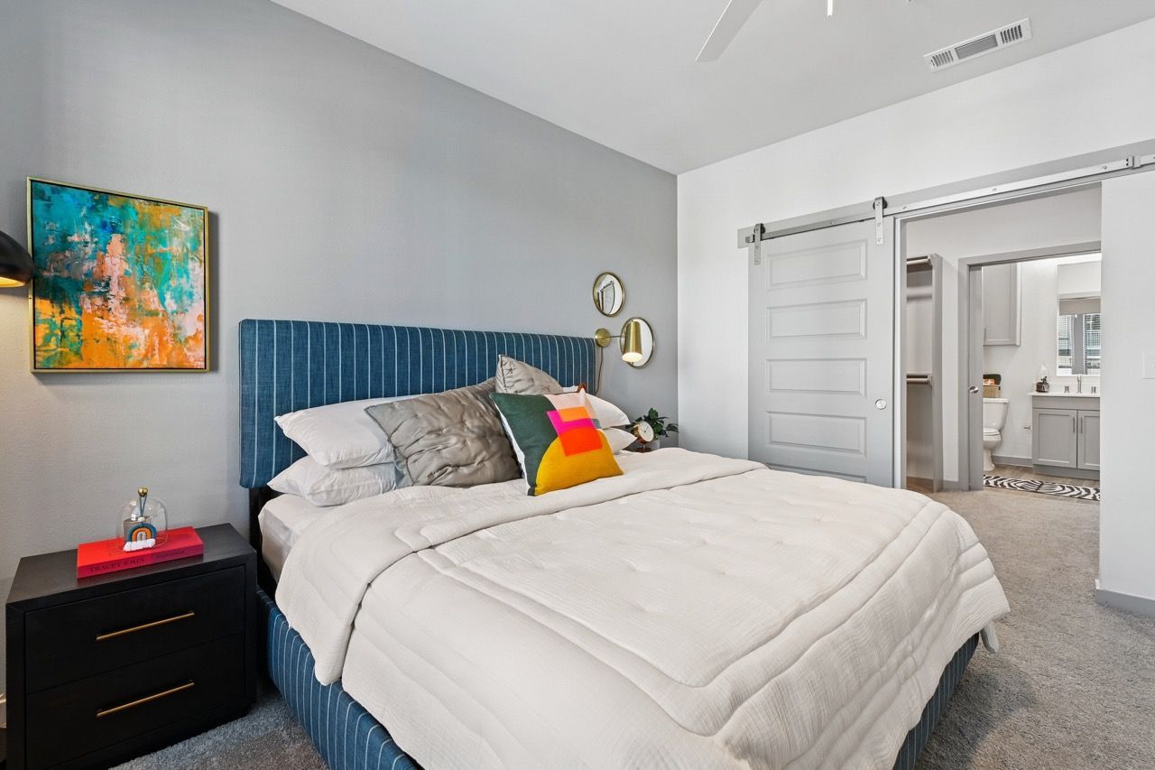 Bedroom with blue upholstered bed, white bedding, nightstand, and doorway to bathroom  at Ennova Fulshear in Fulshear, TX.