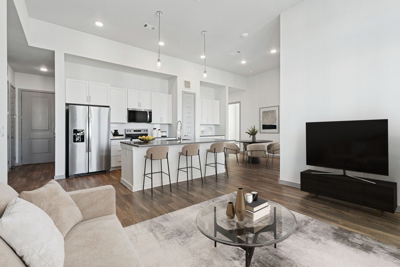 Open-concept apartment living area with a kitchen island, white cabinets, stainless appliances, and a TV  at Ennova Fulshear in Fulshear, TX.