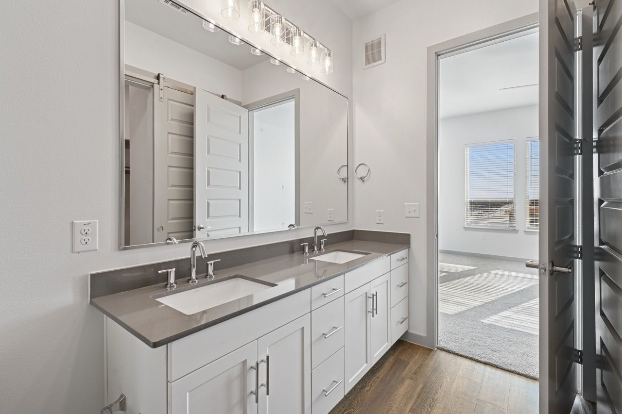 Two-sink bathroom vanity with gray countertop, large mirror, and doorway to a sunlit bedroom  at Ennova Fulshear in Fulshear, TX.