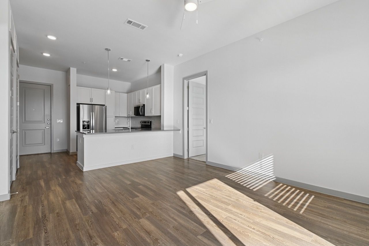 Open-concept apartment living area with white kitchen cabinets, stainless steel appliances, and hardwood floors  at Ennova Fulshear in Fulshear, TX.