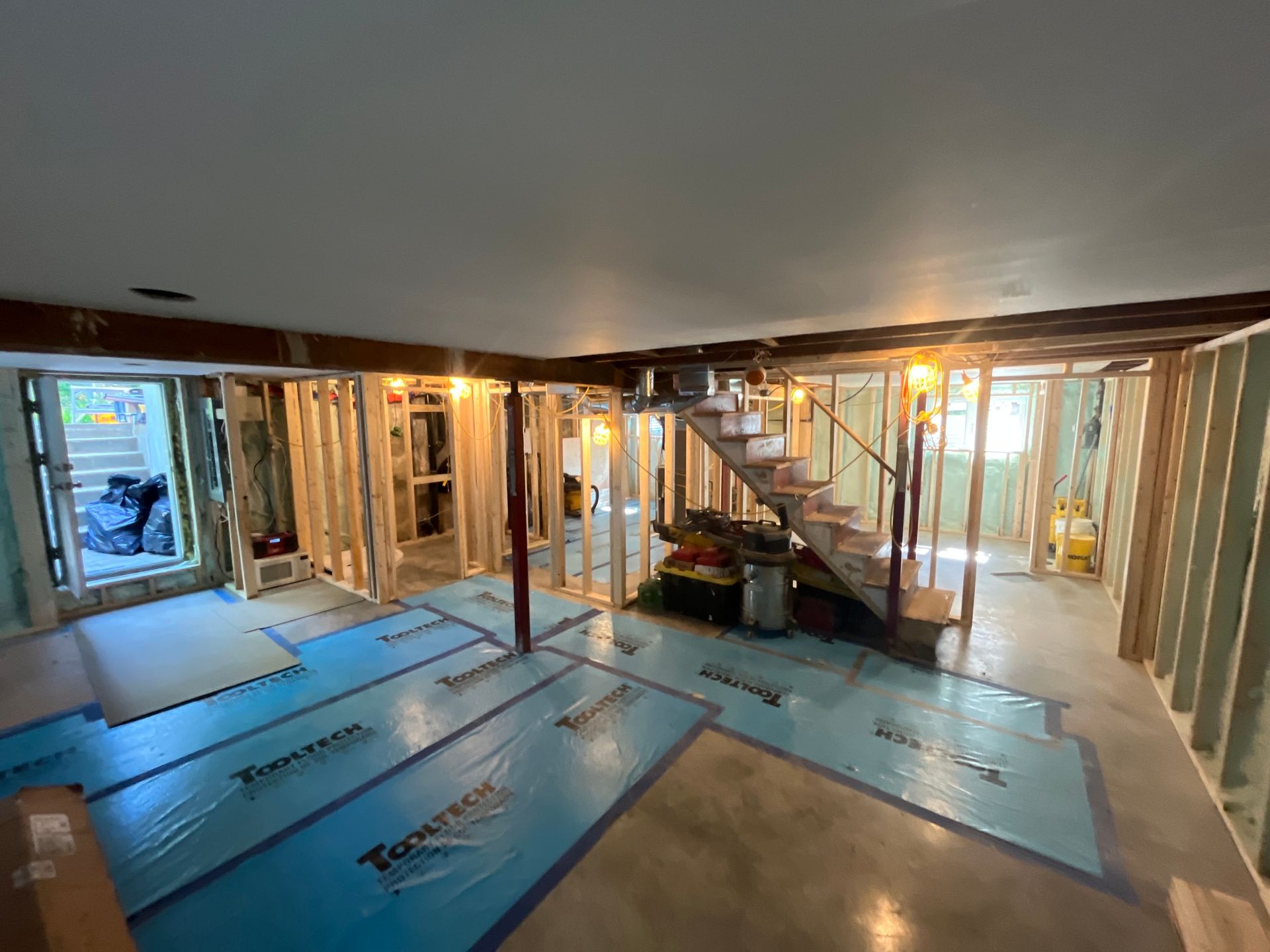 Sous-sol en construction avec charpente en bois, pare-vapeur bleu et escalier. L'espace est éclairé par des plafonniers.