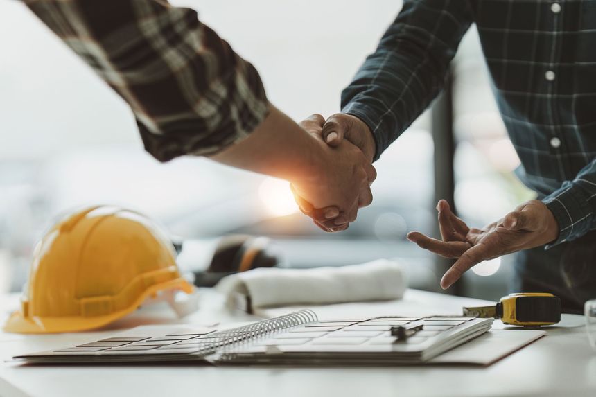 Deux ouvriers du bâtiment se serrant la main au-dessus d'un bureau avec des plans, un casque jaune et des outils.