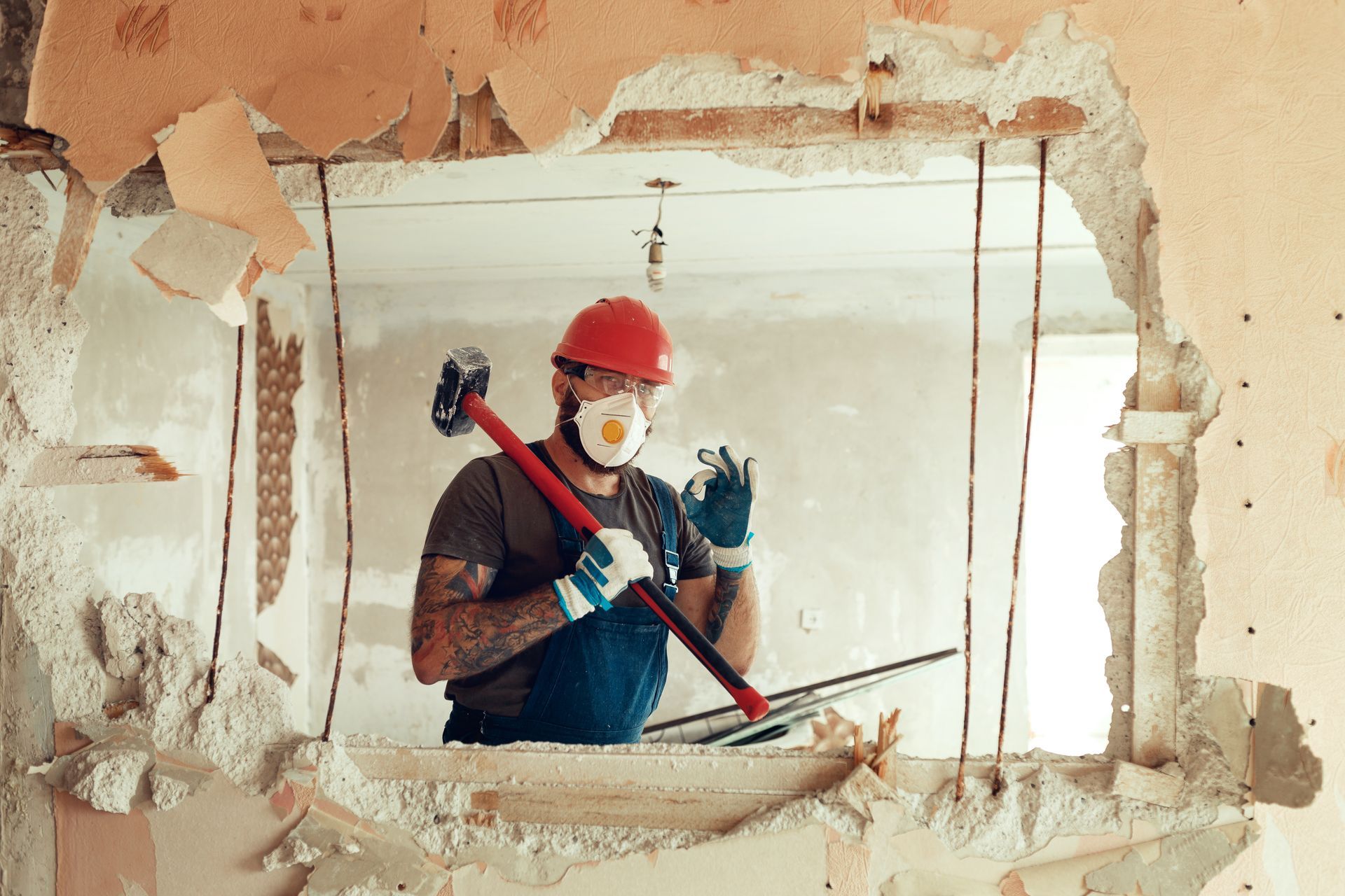 Un ouvrier du bâtiment portant un casque et un masque, brandissant une masse, perce un mur.
