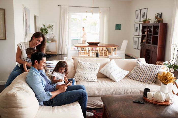 Famille dans un salon lumineux : père sur le canapé utilisant une tablette, mère avec deux enfants, salle à manger en arrière-plan.