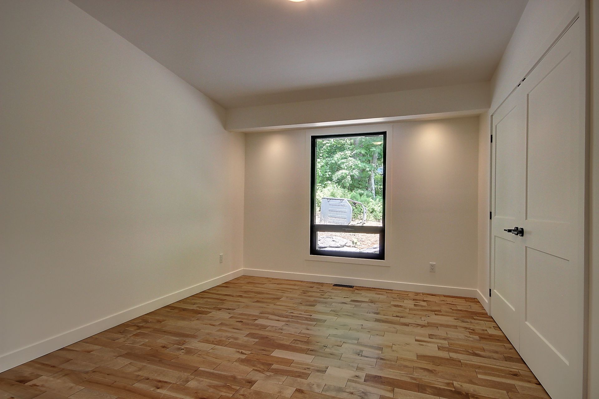 Pièce vide avec parquet, murs blancs, une fenêtre et une porte blanche.
