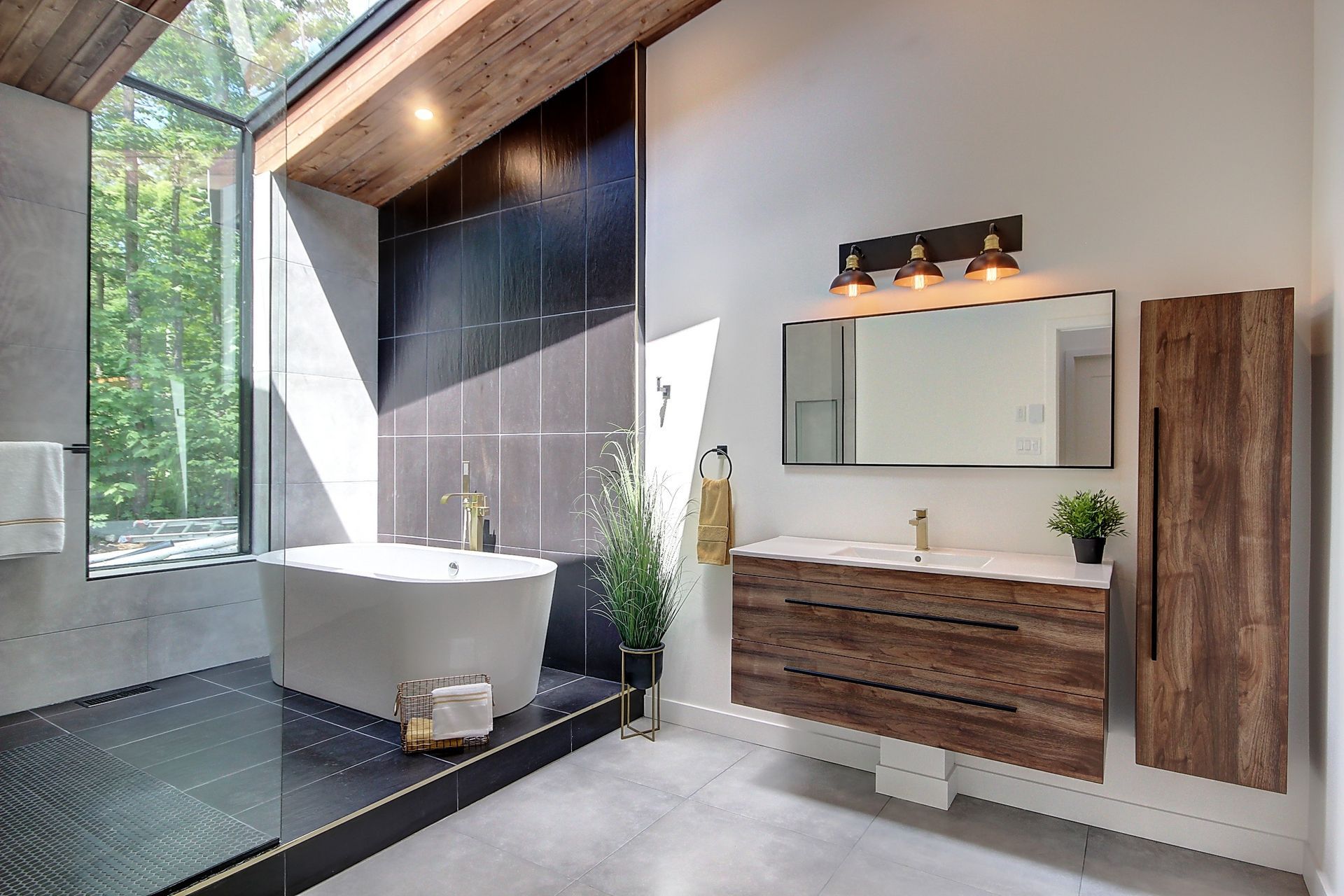 Salle de bain moderne avec baignoire îlot, meuble-lavabo en bois et grande fenêtre donnant sur les arbres. Les murs sont gris, avec du carrelage noir autour de la baignoire.