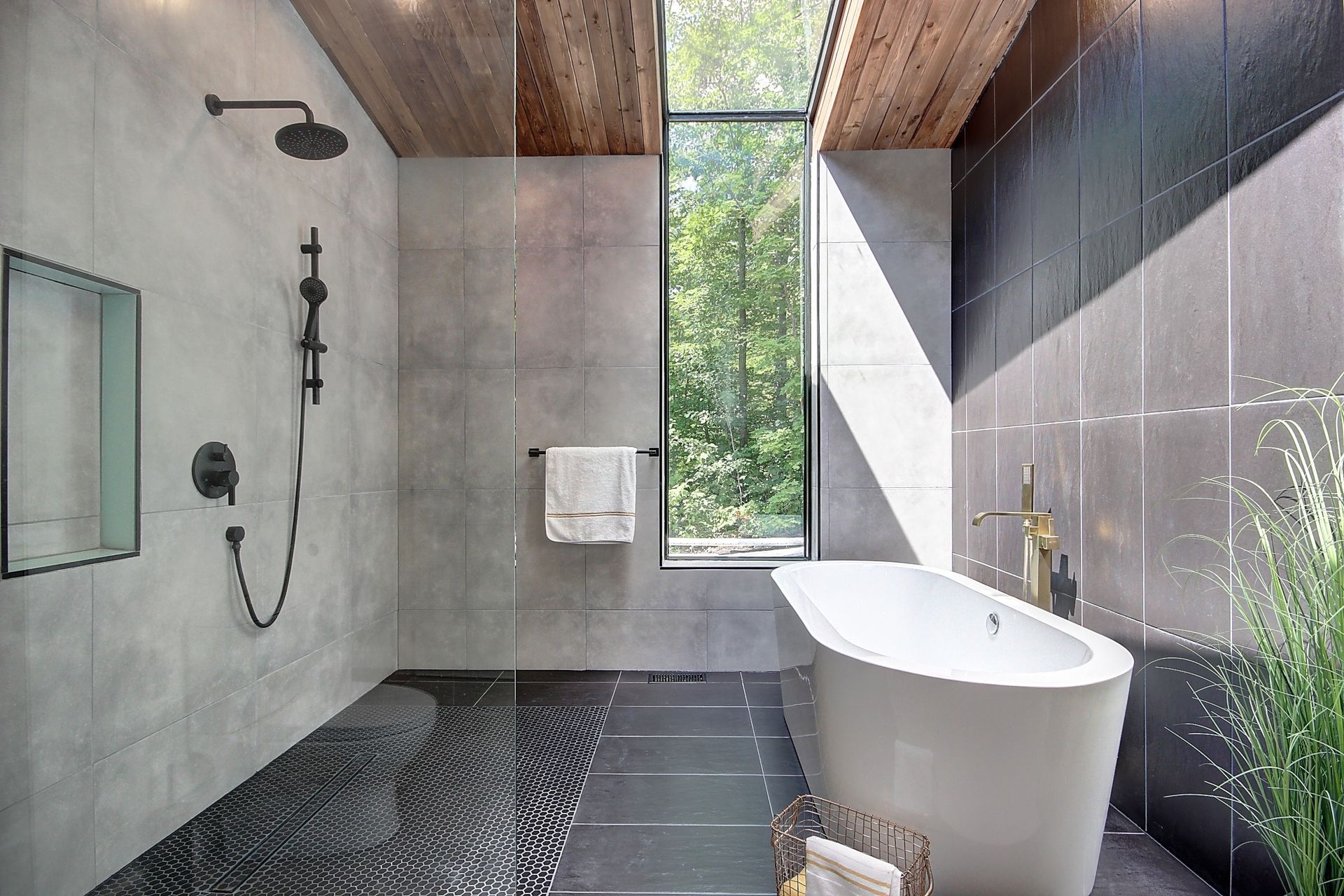 Salle de bain moderne avec baignoire îlot, douche et grande fenêtre donnant sur la verdure. Murs carrelés gris et plafond en bois.