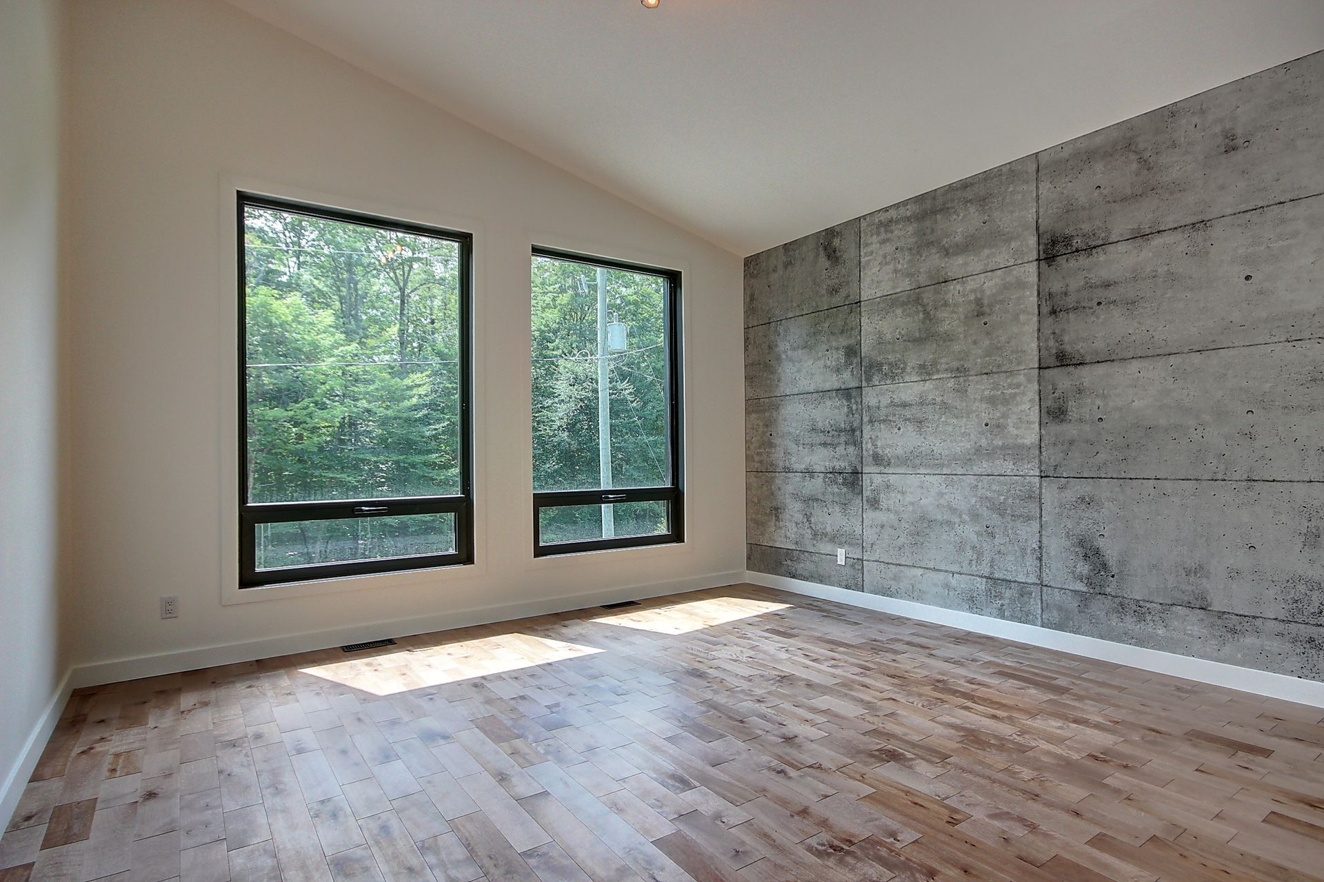 Une pièce vide avec du parquet, un mur en béton et deux grandes fenêtres avec vue sur les arbres.