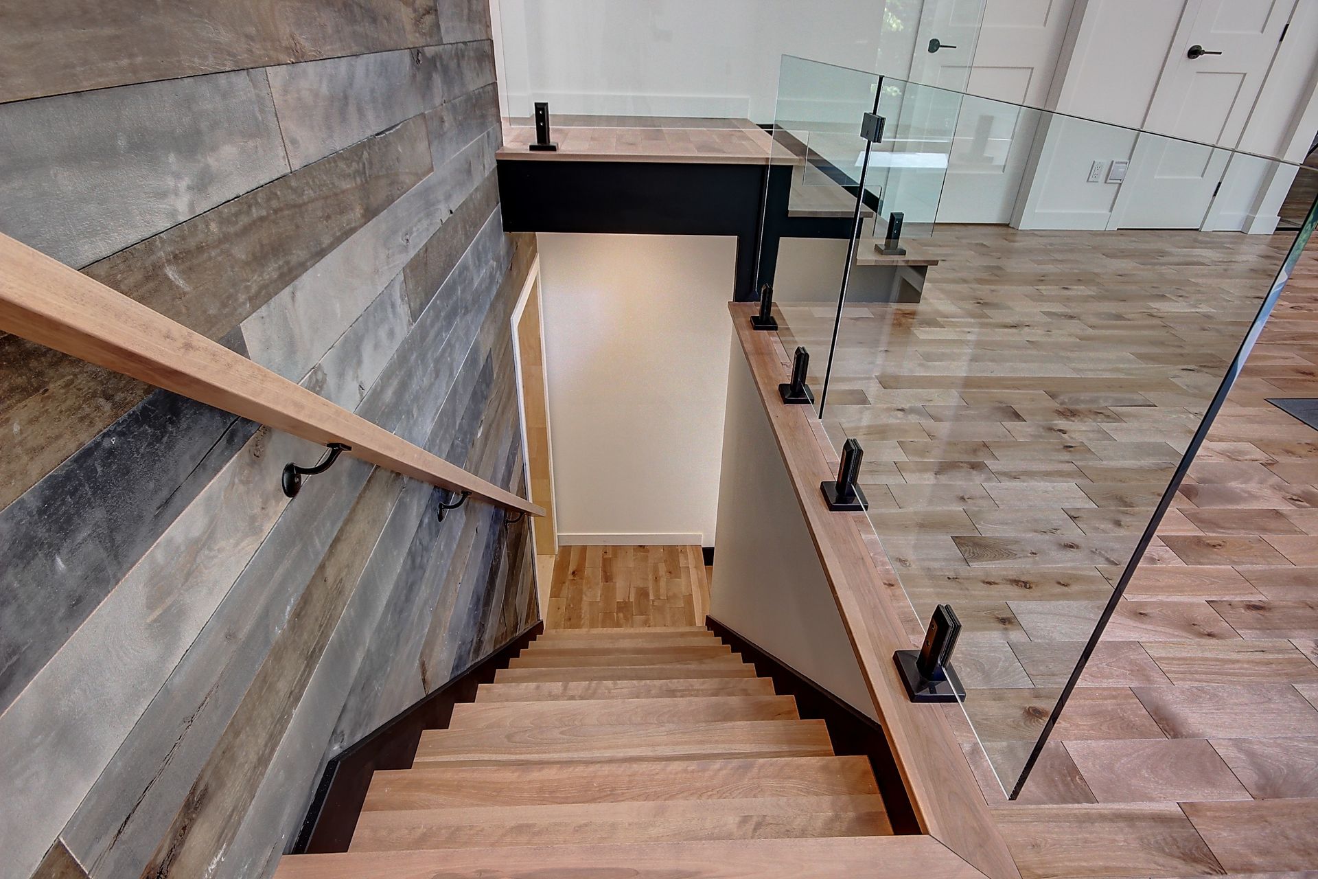 Escalier en bois menant vers le bas. Un mur lambrissé se trouve à gauche, et une rampe en verre à droite.