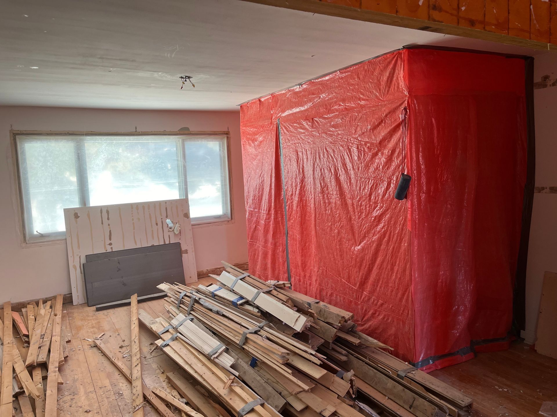 Pièce intérieure en cours de rénovation avec une barrière en plastique rouge. Des débris et du bois encombrent le parquet près d'une fenêtre.