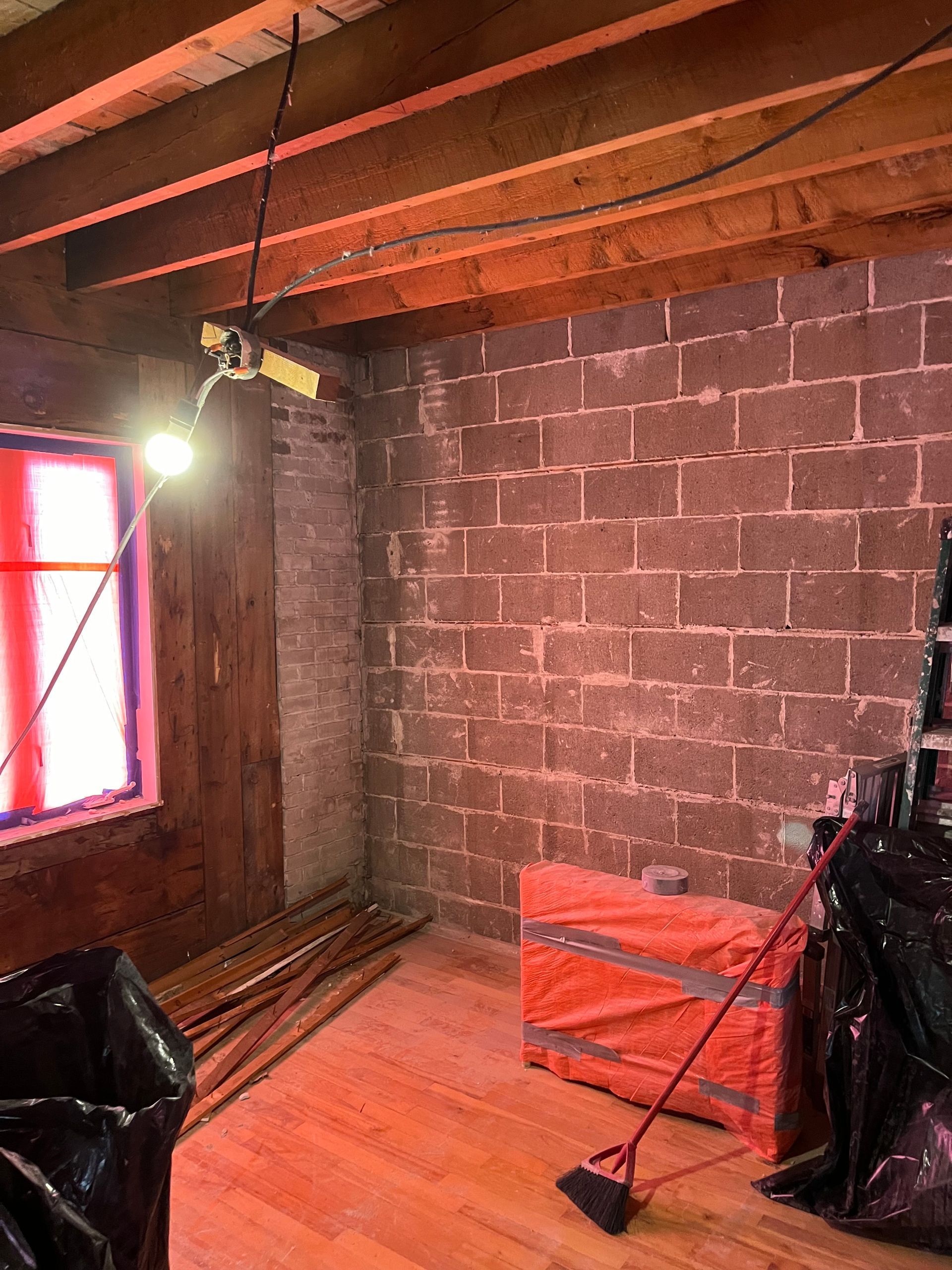 Une pièce avec un mur de briques, des poutres en bois et une fenêtre. Des matériaux de construction sont éparpillés au sol.