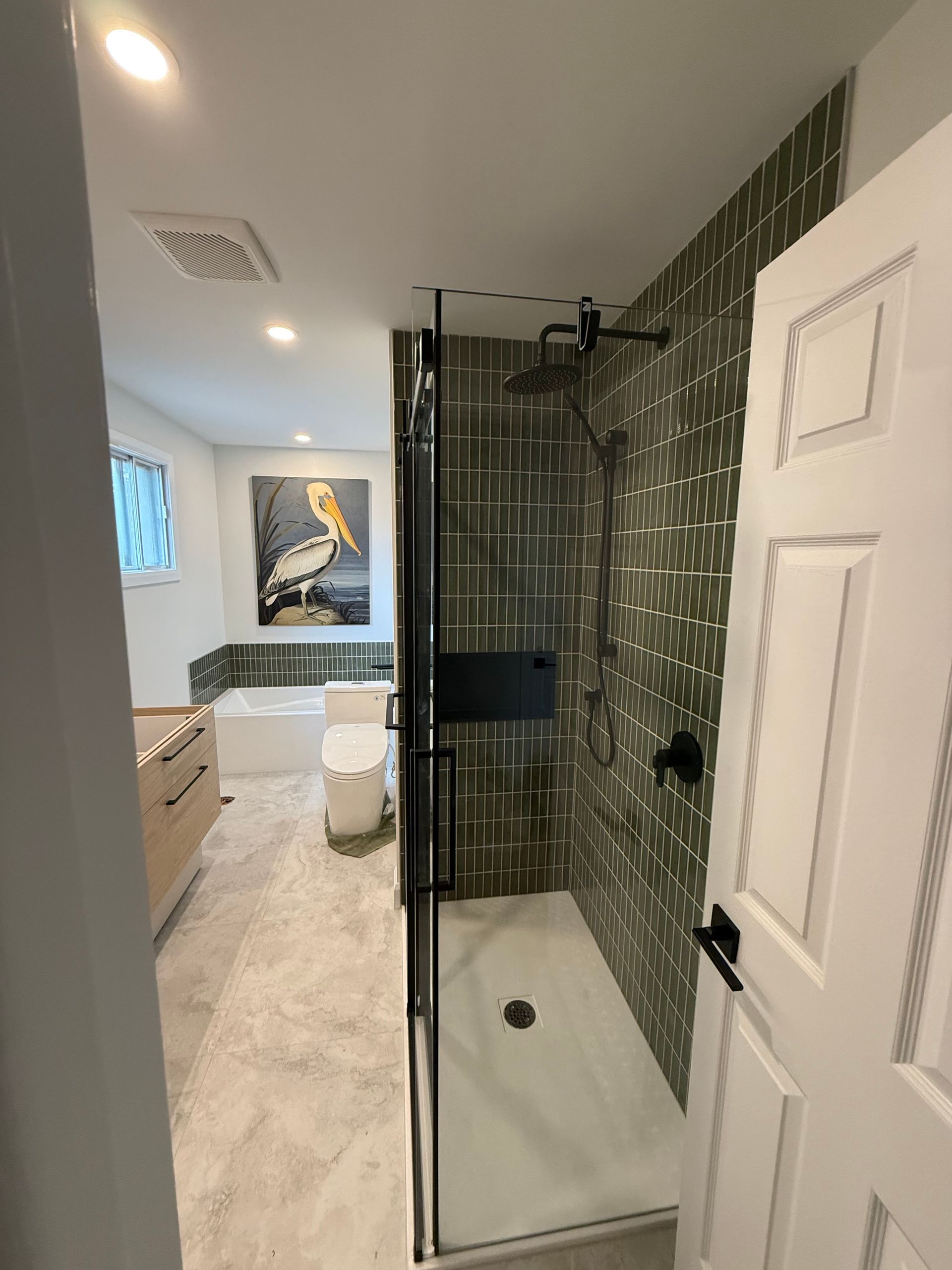 Salle de bain avec douche carrelée verte, porte vitrée, robinetterie noire et œuvre d'art encadrée représentant un oiseau. On aperçoit un meuble-lavabo et des toilettes de couleur claire.