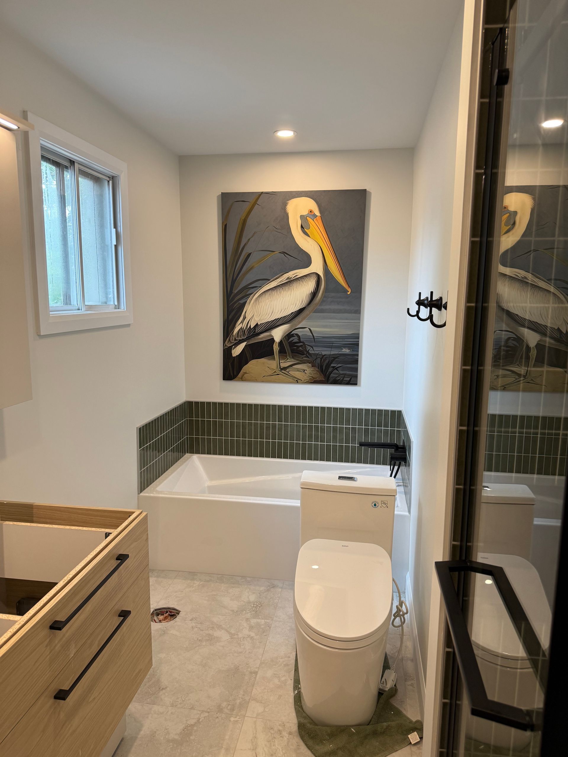 Une salle de bain avec toilettes blanches, baignoire et un tableau représentant un pélican. Les murs sont blancs avec des touches de carrelage vert et une petite fenêtre.
