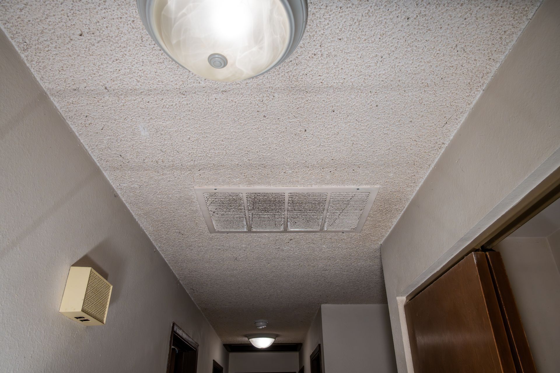 Vue en contre-plongée du plafond d'un couloir avec luminaire et bouche d'aération. Le plafond présente une finition texturée.