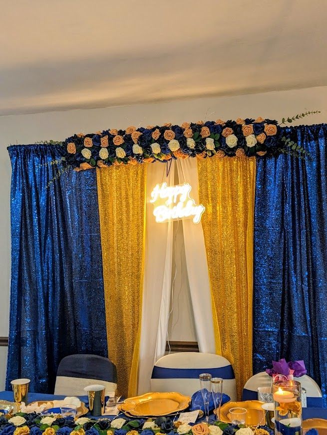 Decorated banquet table with blue and gold drapes, flowers, chairs, and a lit neon sign backdrop