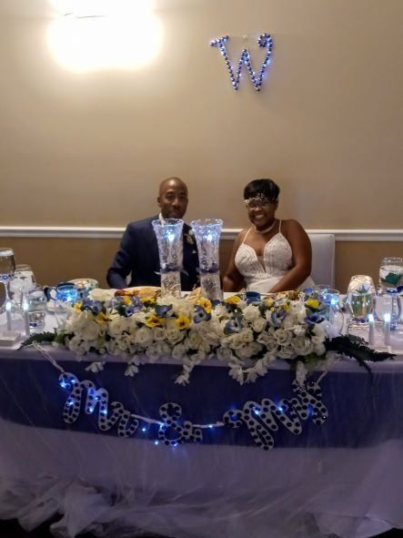 Couple seated at a decorated wedding reception table with blue and white floral centerpieces and a monogrammed backdrop