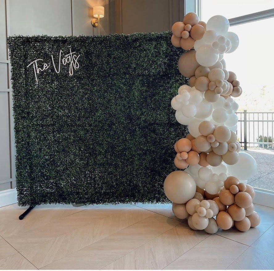 Green hedge photo backdrop with beige, white, and gold balloon garland on the right, indoors
