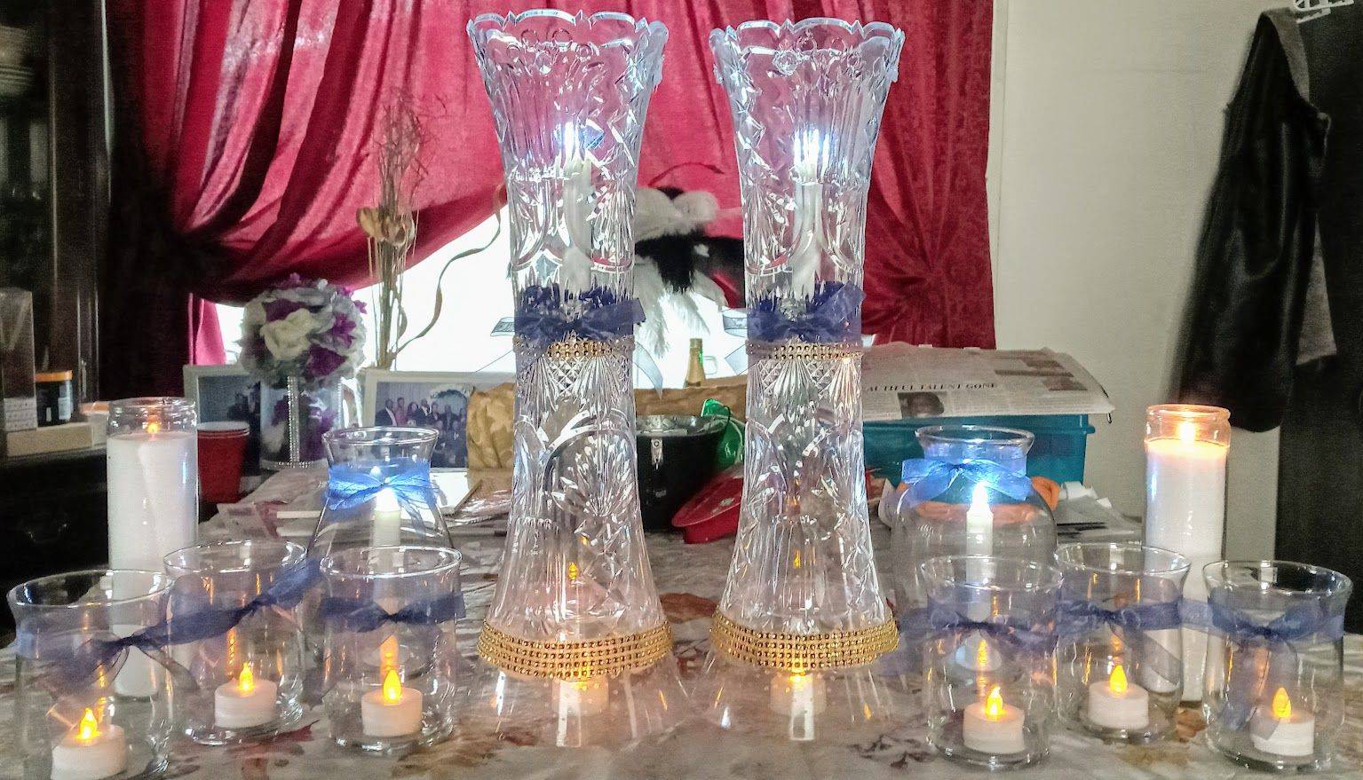 Elegant candlelit table display with two tall crystal vases, glassware, and red drapes in the background