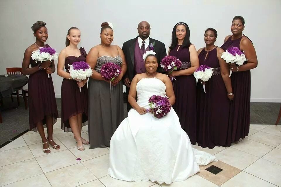 Wedding party posing indoors; bride in white gown seated with bouquet, attendants in burgundy dresses and gray suit.