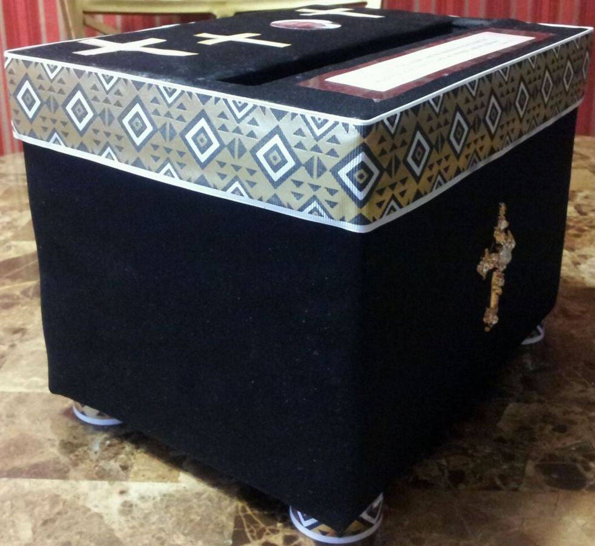 Black box with patterned lid and small cross ornament, resting on wheels or casters.