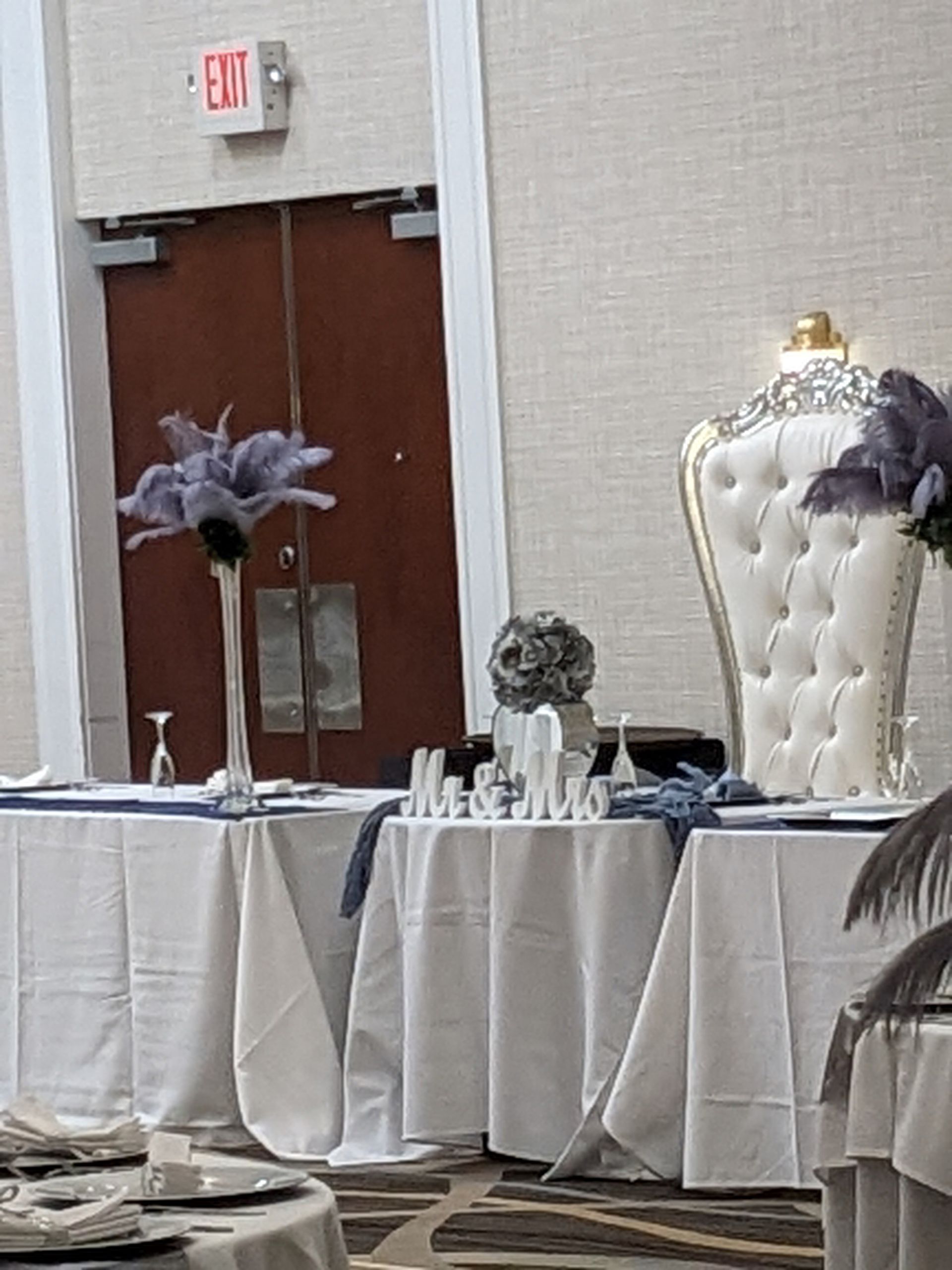 Decorated event table with white linens, purple floral arrangements, and a tall tufted chair in a banquet hall