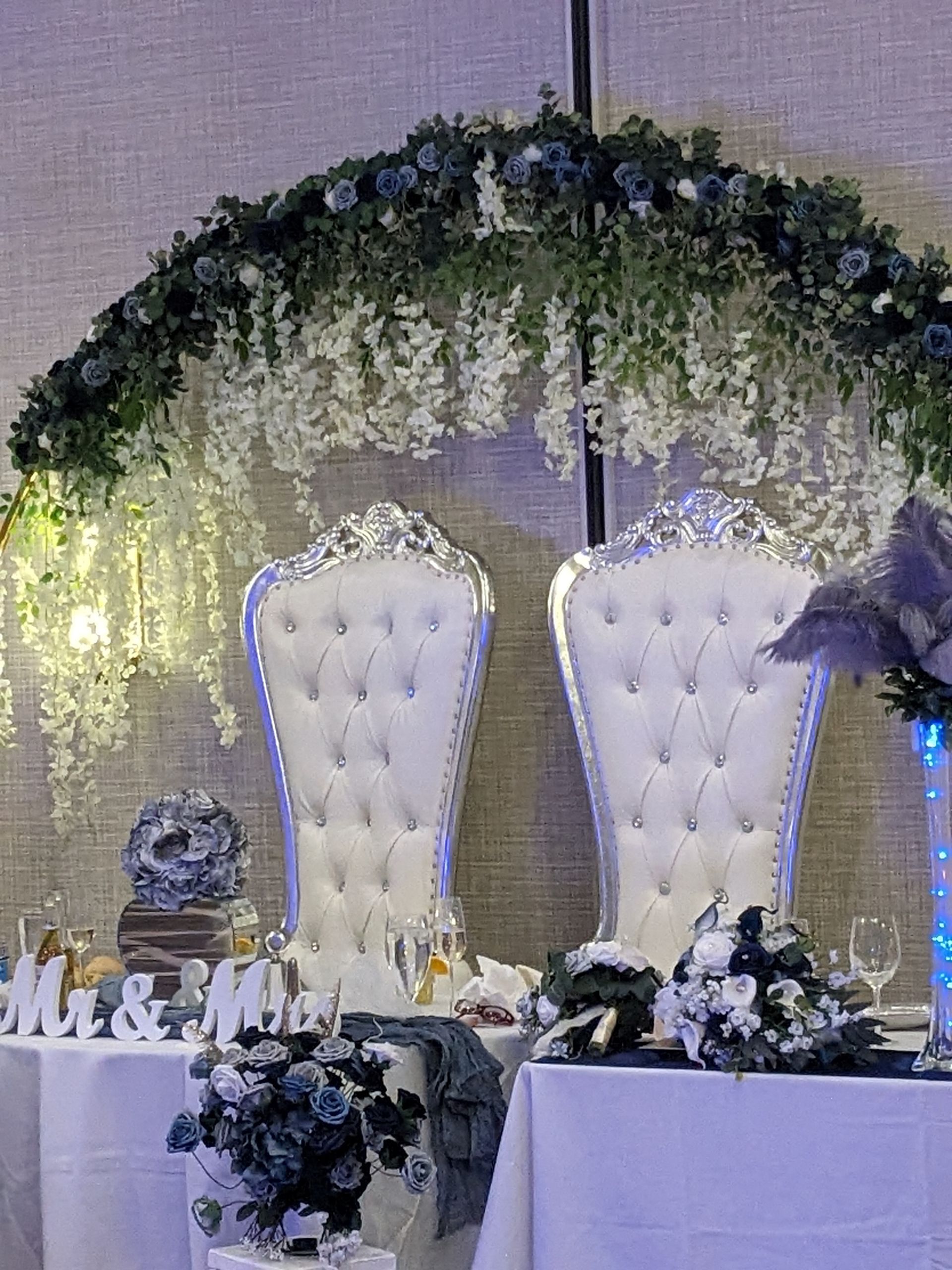 Wedding stage with two white tufted chairs under a floral arch and draped greenery, decorated with candles and white flowers