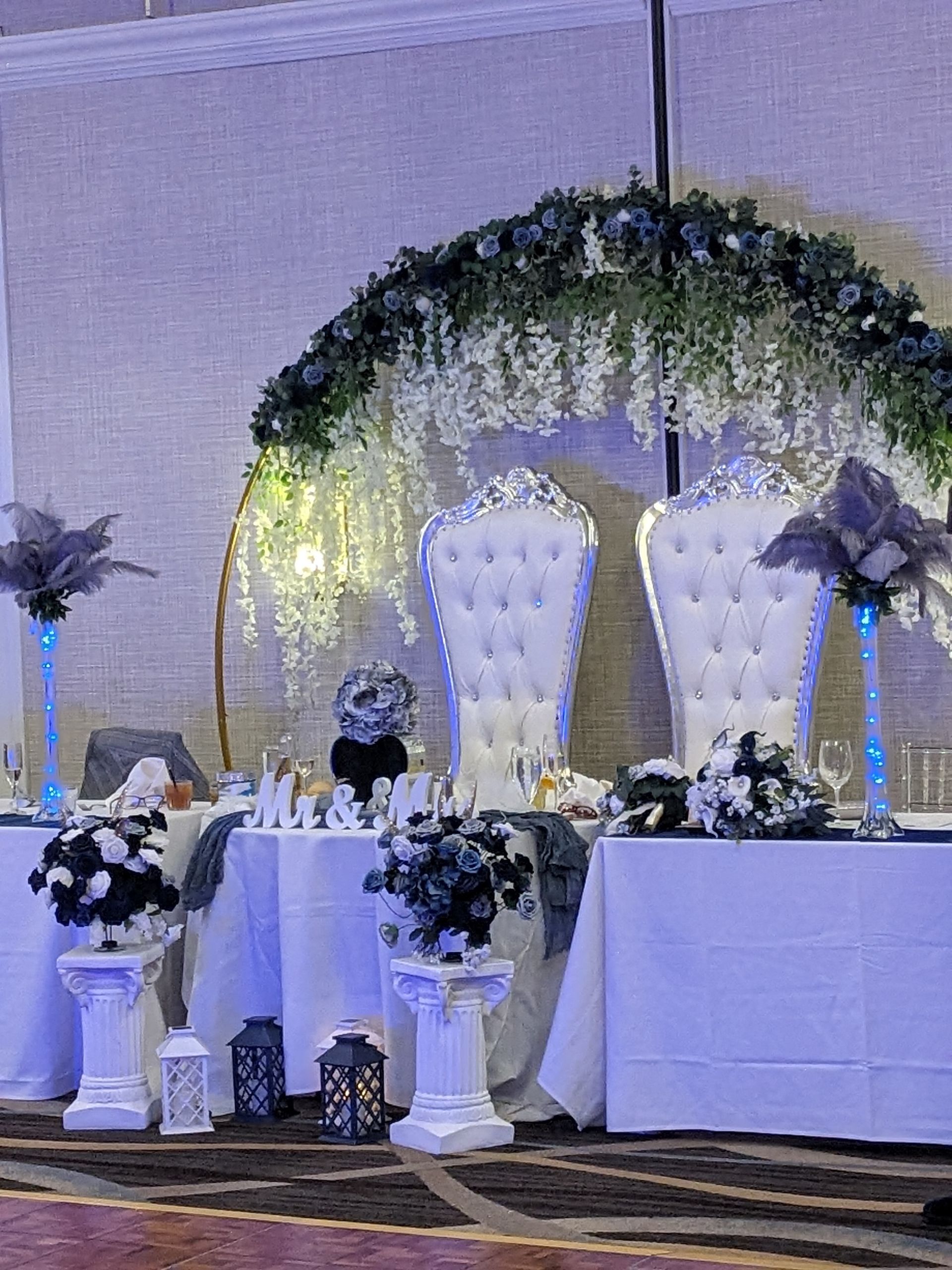Decorated wedding stage with two white throne chairs, floral arch, and candlelit table draped in white and blue flowers