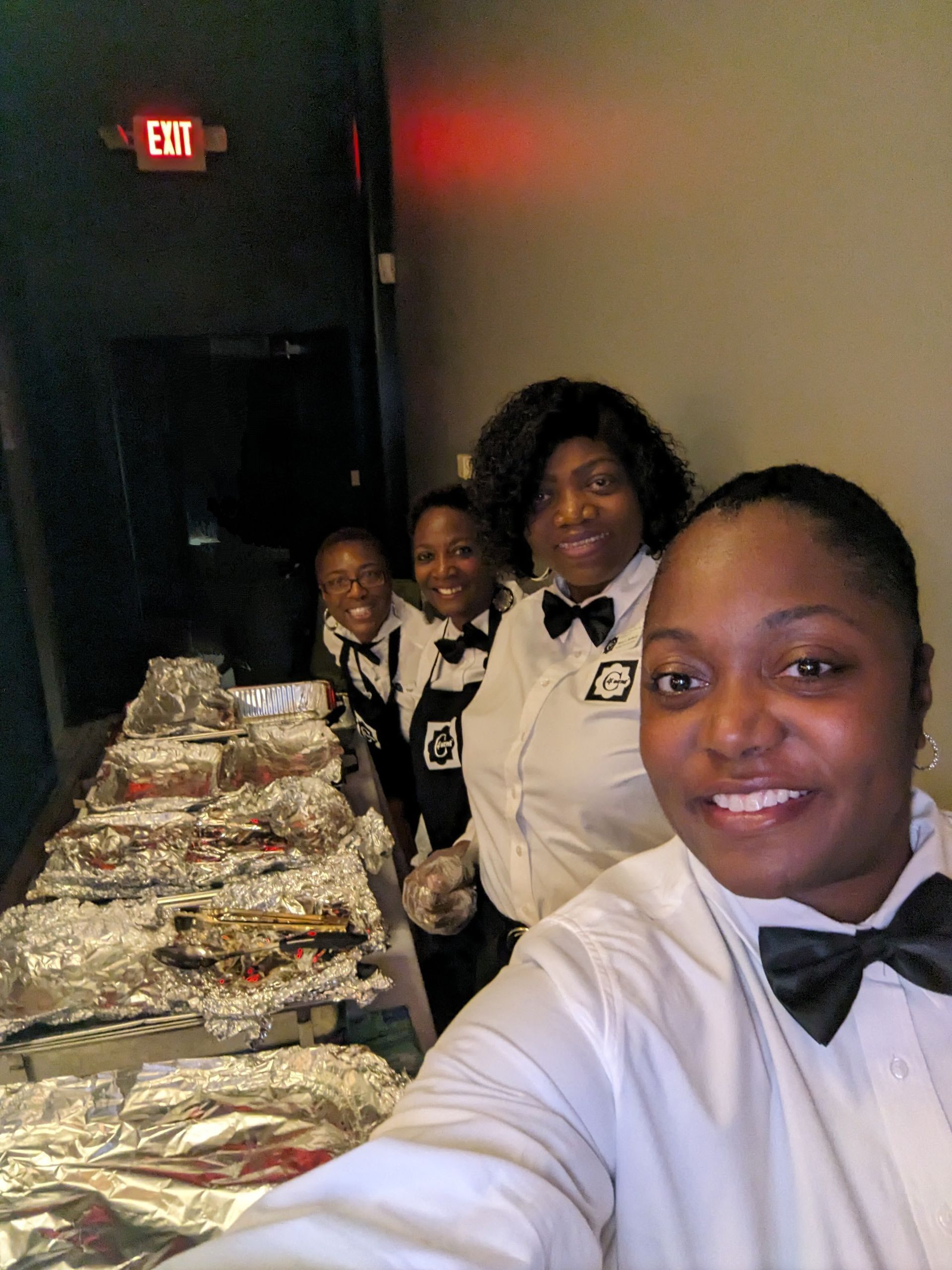 Four people in white shirts and black bow ties pose beside a foil-covered buffet table in a dim hallway.