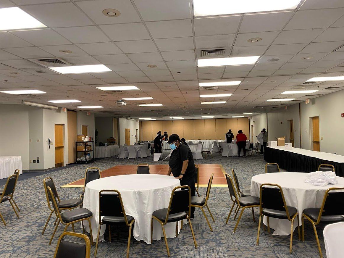 Large banquet hall with round tables, chairs, and a few people near the back wall.