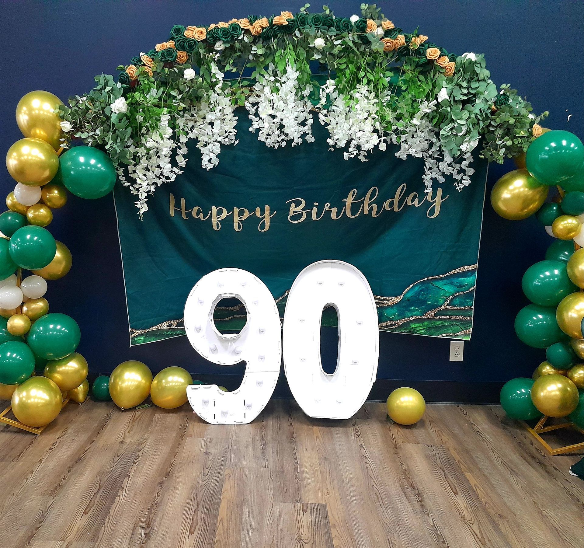 90th birthday backdrop with green-gold balloons, floral arch, and large white “90” numbers