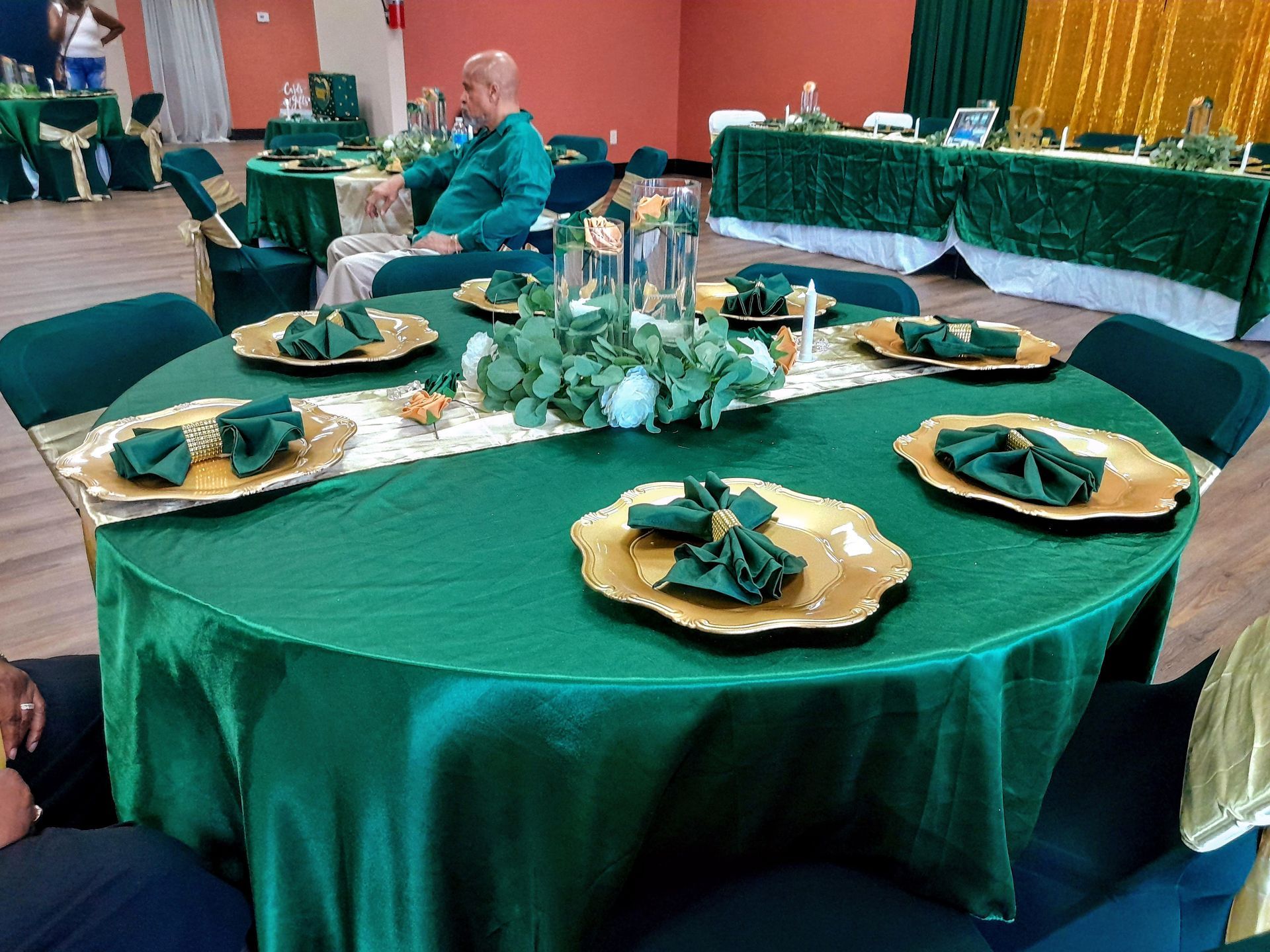 Round banquet table with green tablecloth, gold plates, folded napkins, and floral centerpiece in a decorated hall