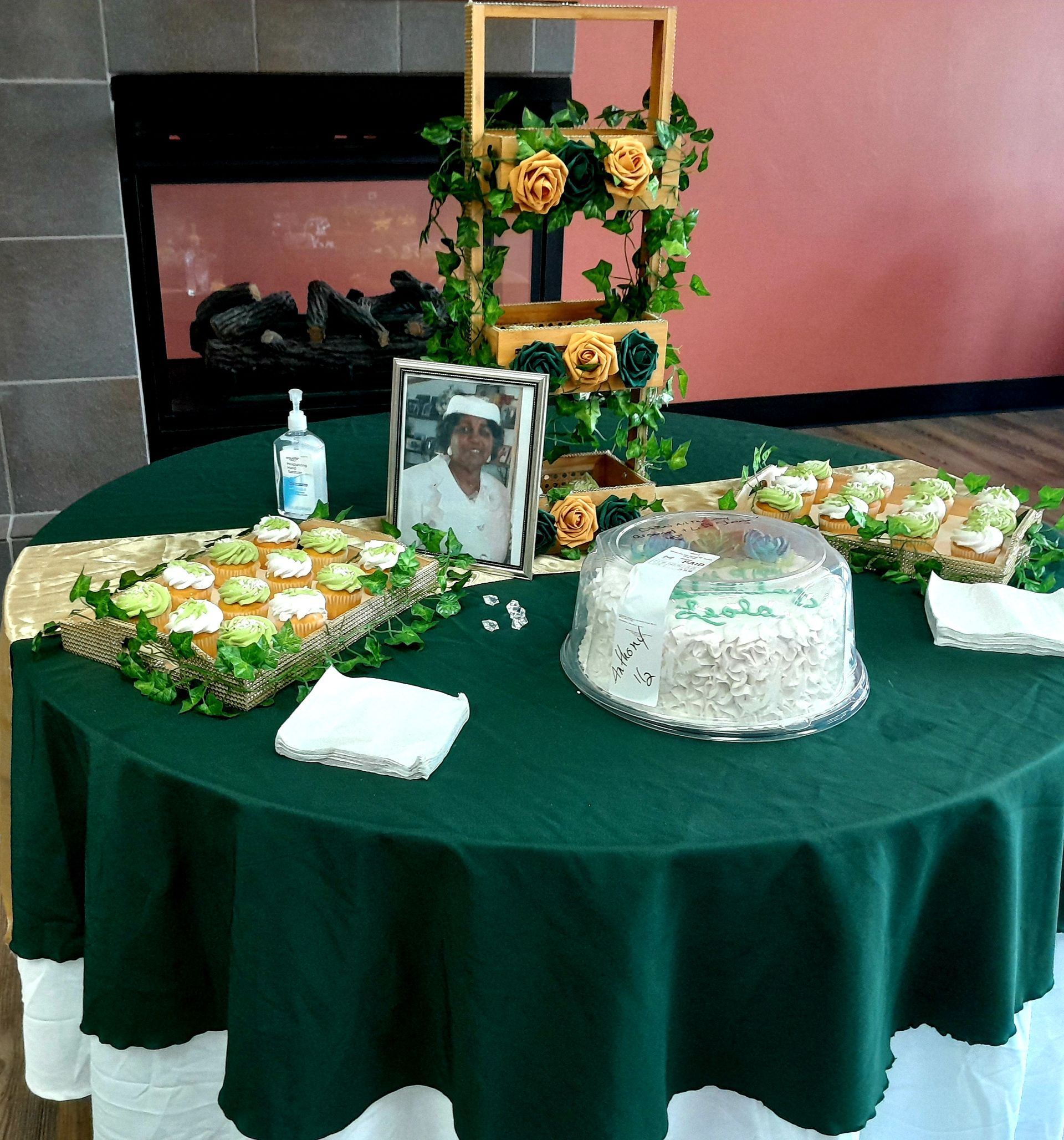 Green-covered memorial table with framed photo, flowers, candles, and a decorated cake-shaped centerpiece