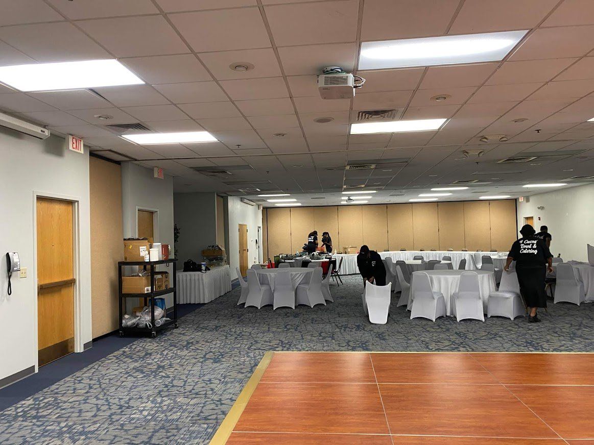 Large banquet room with white tablecloths, a carpeted floor, and a few people standing near the back.