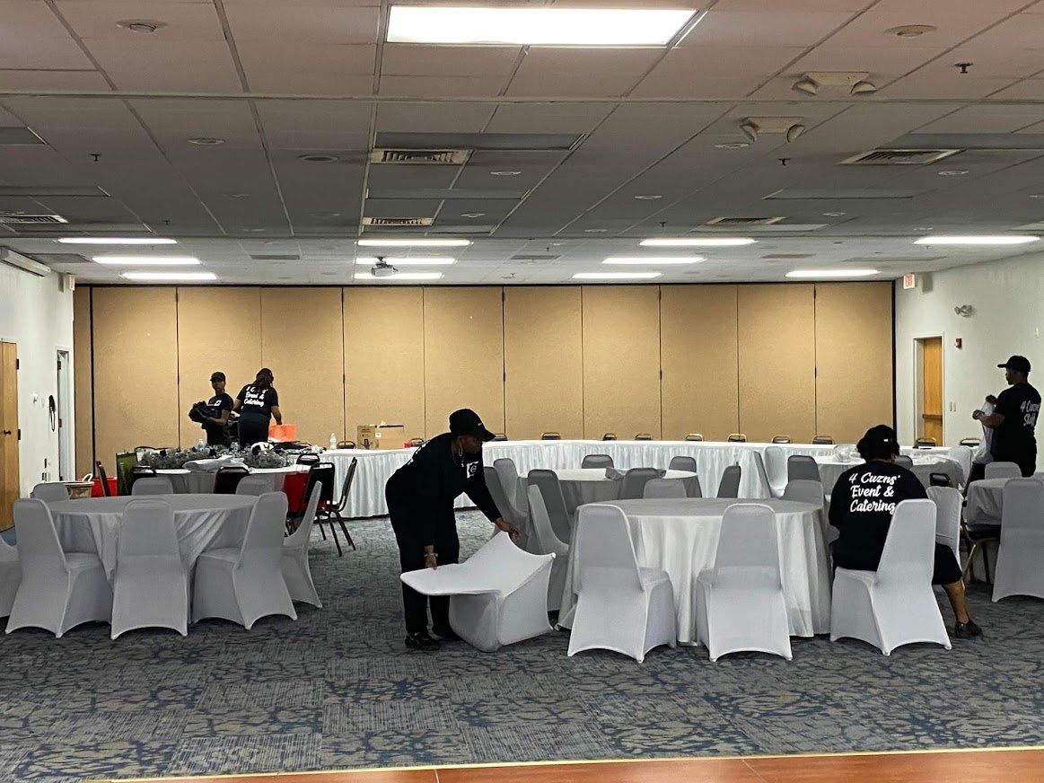 Banquet hall with round tables and white chair covers being set up by staff near a stage area