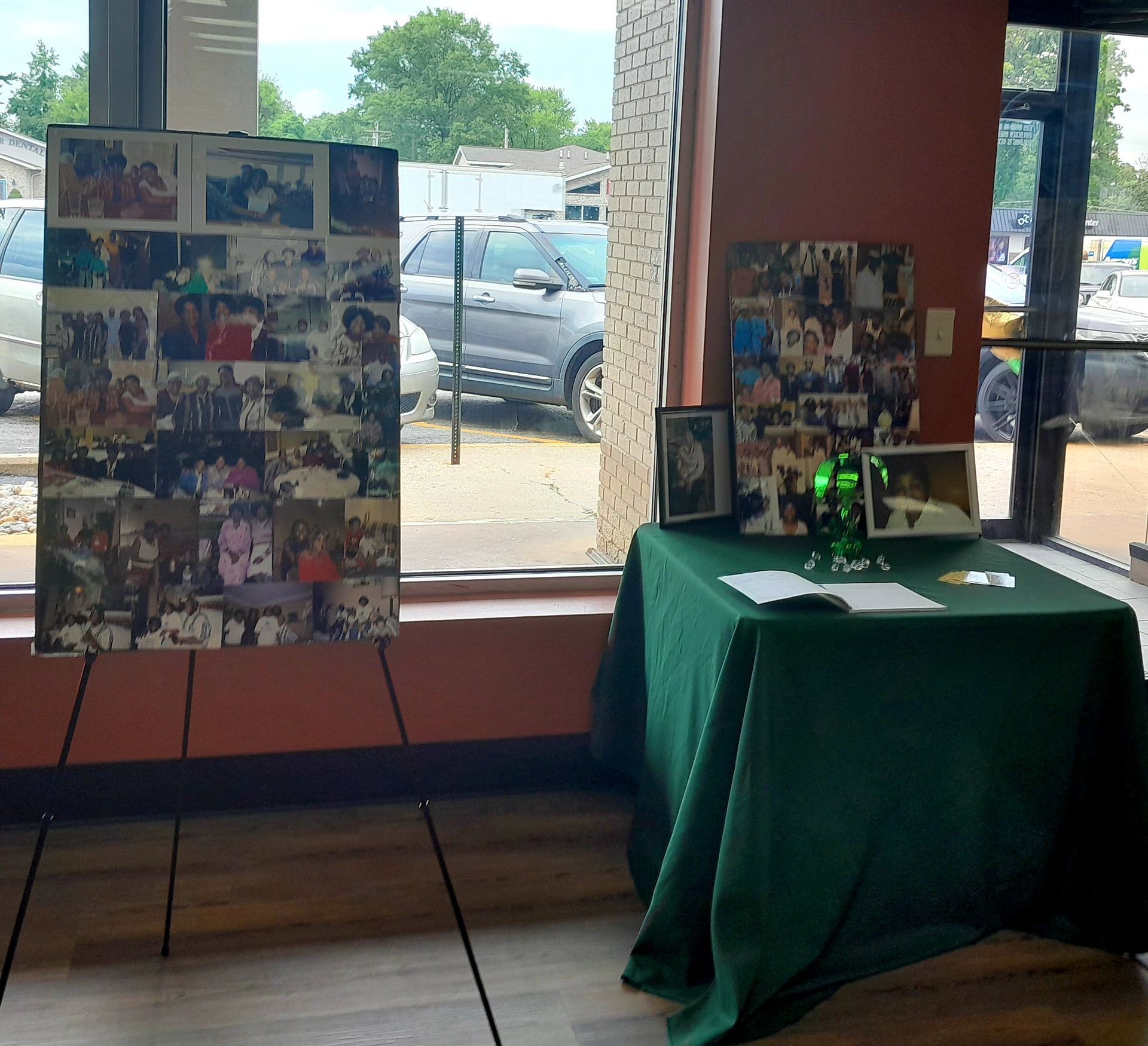 Photo collage boards and a green-draped display table in a windowed room overlooking parked cars.
