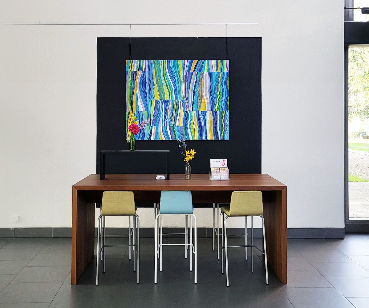 Een tafel met stoelen voor een zwarte muur die is versierd met een kleurrijk, abstract Jo & Josie schilderij.