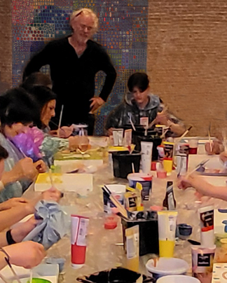 Onder toeziend oog van Jo Debije zit een groep mensen aan een lange tafel te schilderen in een atelier.