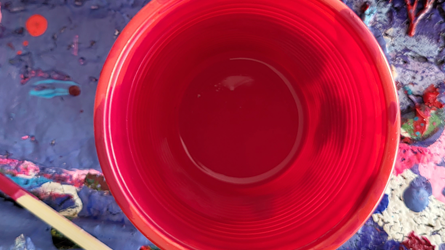 Een bovenaanzicht van een lege rode plastic beker op een oppervlak dat bedekt is met vlekken blauwe, roze en witte verf.