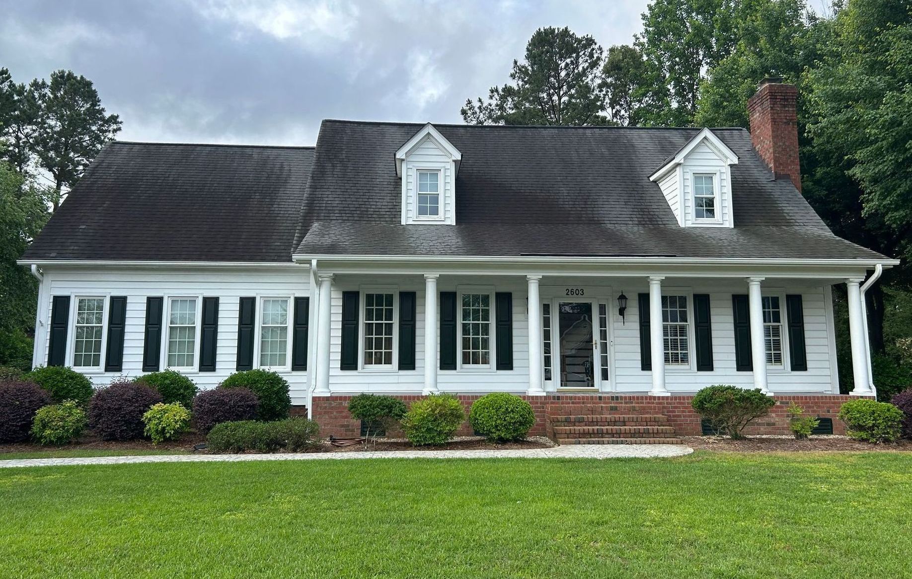 White house with black shutters, dormers, and porch, red brick chimney, and green lawn.