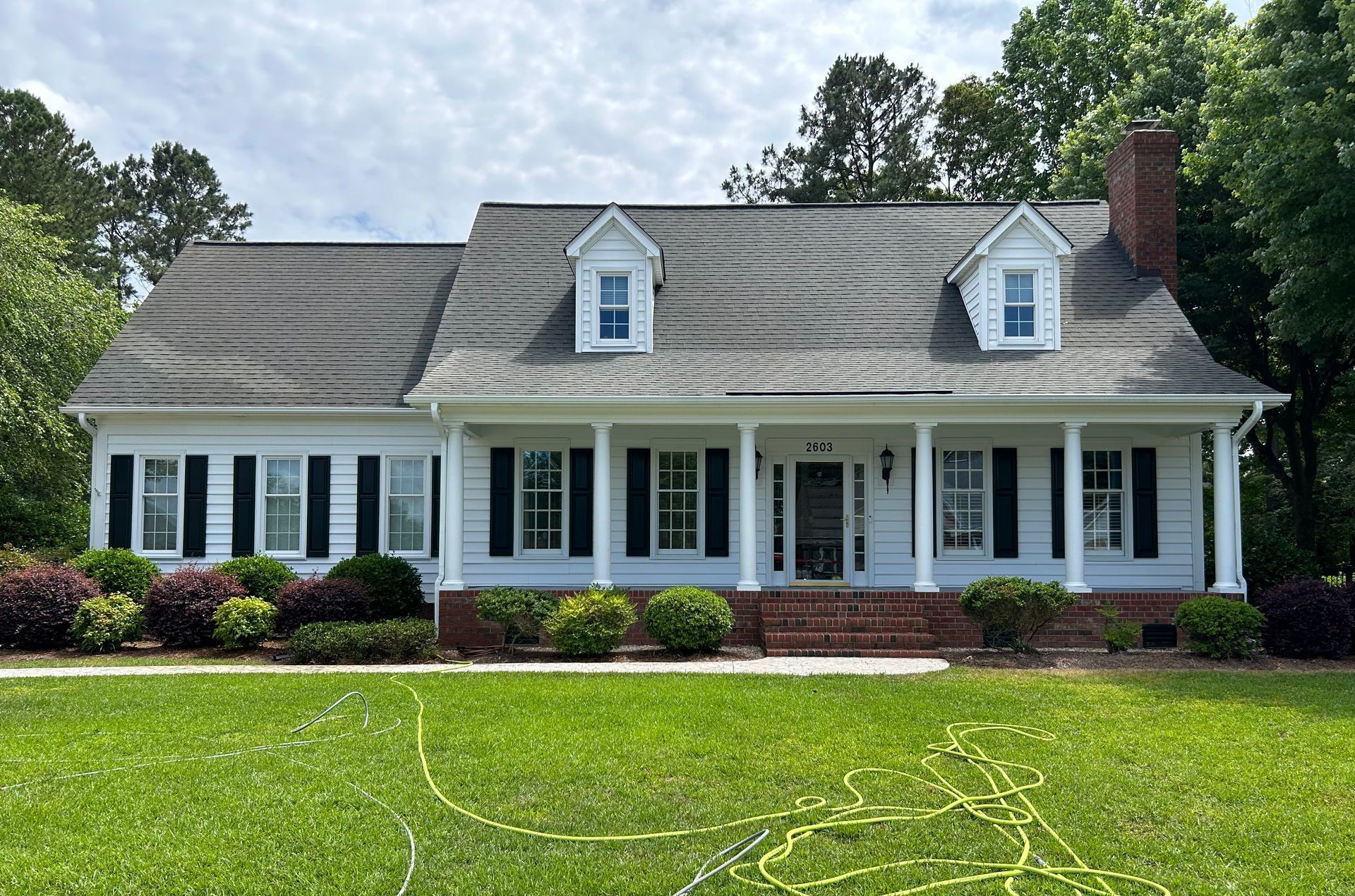 White house with black shutters, dormers, and a brick chimney, fronted by green lawn and bushes.